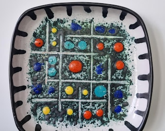 May include: Square ceramic plate with a black rim and a white border featuring black, vertical, and horizontal lines. The center has a green speckled background with colorful dots in orange, blue, yellow, and turquoise.