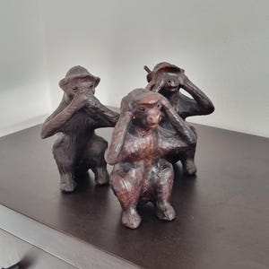 May include: Three brown monkey figurines in the "See no evil, hear no evil, speak no evil" pose. The sculptures are on a dark brown surface, with a white wall in the background. The monkeys are detailed with textured fur and facial features.