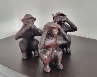 May include: Three brown monkey figurines in the "See no evil, hear no evil, speak no evil" pose. The sculptures are on a dark brown surface, with a white wall in the background. The monkeys are detailed with textured fur and facial features.