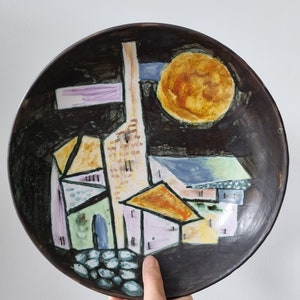 Puede incluir: Un plato de cerámica con fondo negro y un diseño abstracto colorido. El diseño presenta un sol amarillo, una franja púrpura y blanca, y un grupo de edificios en tonos de verde, amarillo, rosa y azul.