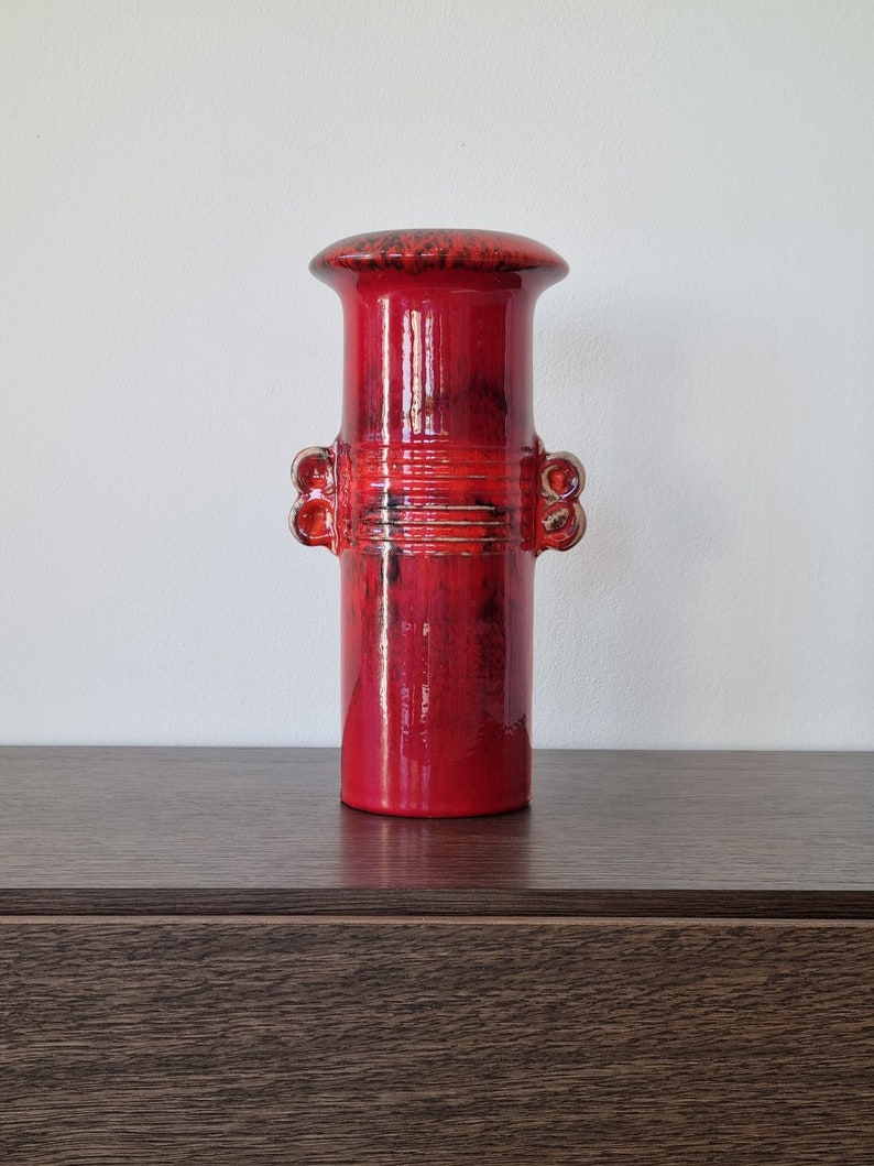 May include: A red ceramic vase with a textured surface and a wide, round opening. The vase has a cylindrical shape with two decorative bands around the middle.