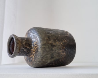 Könnte beinhalten: Eine dunkle, strukturierte Keramikvase mit rundem Körper und zylindrischem Hals. Die Vase hat ein gesprenkeltes Aussehen mit Grautönen, Braun und Gold. Das Innere des Halses ist rotbraun. Die Vase liegt auf der Seite.