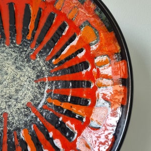 Puede incluir: Un plato de cerámica con un diseño llamativo. El plato presenta un borde negro y un patrón central de formas rojas, naranjas y negras que irradian desde un centro texturizado blanquecino. El diseño tiene una estética moderna de mediados de siglo.