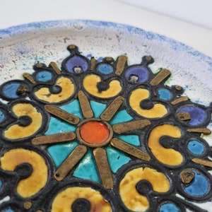 Op de afbeelding: Een keramisch bord met een kleurrijk, geometrisch ontwerp. Het midden van het bord heeft een stervormig patroon met een rood en oranje midden, omringd door gele, blauwe en turquoise tegels. Het bord heeft een witte achtergrond met een blauwe rand.