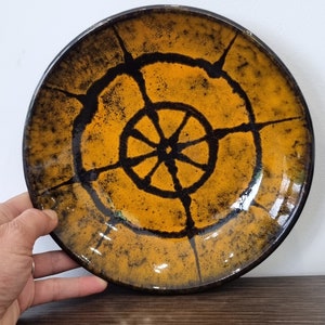Op de afbeelding: Een rond keramiek bord met een zwart en geel ontwerp. Het bord heeft een zwart midden met een stralend patroon van lijnen en een gele buitenring.