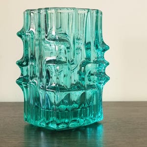 May include: A translucent teal glass vase featuring a thick, molded geometric pattern with vertical ridges and prominent, pointed protrusions extending from the sides. The vessel has a rectangular body with a clear, reflective surface.
