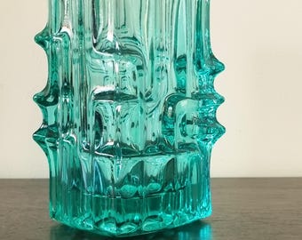 May include: A translucent teal glass vase featuring a thick, molded geometric pattern with vertical ridges and prominent, pointed protrusions extending from the sides. The vessel has a rectangular body with a clear, reflective surface.