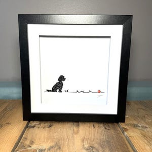 May include: Framed art print with a black frame, depicting a black silhouette of a dog seated and gazing at a small orange ball. The dog is on a line representing grass, set against a white backdrop.