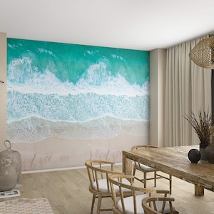Può includere: Una sala da pranzo con un murale a parete che mostra una vista aerea di onde oceaniche che si infrangono su una spiaggia sabbiosa. La stanza ha un tavolo in legno con sedie, un lampadario intrecciato e un arazzo da parete intrecciato.