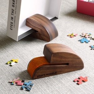 May include: Two curved dark wood puzzle holders with a smooth finish. One holder supports a white puzzle box labeled EYES IN THE GARDEN by LISA CONGOON. The other holder features an orange felt insert and the text POP'S PUZZLE printed on the side.