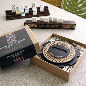 May include: A boxed domino train game set with a wooden holder and dominoes. The box is open, revealing the game pieces and a circular hub. The text "DOMINO TRAIN GAME MAINLINE EDITION" is visible on the box.