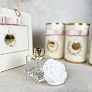 May include: A clear, square glass diffuser bottle with a gold cap, a white flower, and a gift bag with a pink ribbon. The bottle is next to a white flower and several cylindrical containers with gold accents and pink ribbons. The text "Rose in Paradise" is on the bottle.