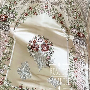 May include: Cream-coloured prayer rug with detailed floral embroidery in red, green, and white. It has a central floral design and decorative borders. The text "ROSE IN PARADISE" is visible.