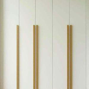 May include: A white wardrobe with four doors and vertical wooden handles. The handles are a light brown color and are rectangular in shape. The wardrobe is set against a light-colored wall and a wooden floor.