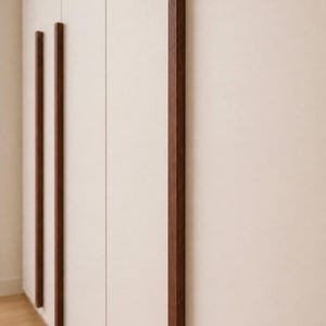 Puede incluir: Tiradores verticales de madera en color marrón oscuro. Los tiradores rectangulares están fijados a una puerta de armario de color claro. El diseño es minimalista y moderno, adecuado para la decoración del hogar. Miden unos 100 cm de largo.