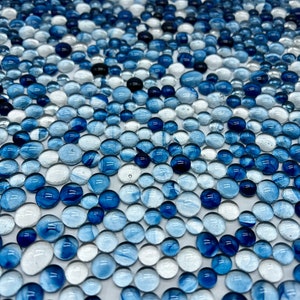May include: An overhead shot of a collection of glass marbles in shades of blue, white, and black. The marbles vary in size and opacity, creating a textured, visually appealing pattern. The marbles are scattered across the surface.