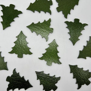 May include: A collection of dark green leather Christmas tree cutouts scattered on a white surface. The cutouts vary in shape, some resembling traditional trees and others more abstract designs. The leather has a textured appearance.