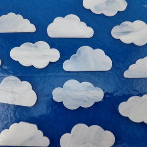 May include: White cloud-shaped decorative pieces against a vibrant blue background. The clouds have a marbled appearance, creating a textured effect. The arrangement suggests a sky-themed design, suitable for crafts or decor.