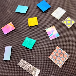 May include: A collection of small, colorful, geometric glass tiles. The tiles are various shapes, including squares and rectangles, and feature a range of colors like blue, yellow, green, and iridescent hues. Some tiles have patterned designs.