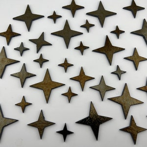 May include: A collection of star-shaped wooden cutouts in varying sizes. The stars have a rustic, weathered appearance with shades of brown and gray. The cutouts are arranged on a white surface.