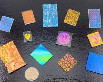 Puede incluir: Una colección de once piezas acrílicas pequeñas, planas, rectangulares y cuadradas con varios patrones, incluyendo un corazón amarillo, un escarabajo rosa y acabados iridiscentes, dispuestas con una moneda para escala.