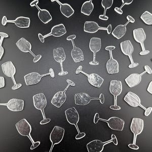 May include: A collection of clear, transparent wine glass cutouts scattered on a black surface. The cutouts vary in orientation, with some upright and others tilted, creating a dynamic arrangement. The glasses have a classic wine glass shape.