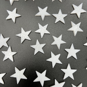 May include: A collection of white star-shaped embellishments scattered on a black surface. The stars have a textured appearance, with some appearing solid and others with a subtle pattern. These decorative stars are suitable for crafts and embellishments.