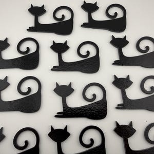 May include: A collection of eleven identical black cat silhouettes arranged in rows. Each cat figure features a pointed head with ears, a long body, and a tail that curls upward into a spiral. The surface of each cat cutout has a textured finish.