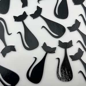 May include: A collection of glossy black cat silhouettes in various poses. The cats have pointed ears, curved tails, and are arranged on a white background. The shapes vary slightly, adding visual interest to the design.