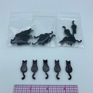 May include: Black cat-shaped embellishments in various poses, some in clear plastic bags, others arranged in a row. A ruler is placed below the cats, providing a size reference. These decorative pieces are ideal for crafting or scrapbooking.