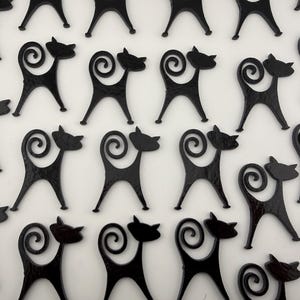 May include: A collection of black cat-shaped silhouettes arranged in a grid pattern. Each cat figure features a smooth, glossy finish, a pointed head, and a long, curled tail that loops upward, creating a repeating decorative motif across the frame.