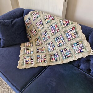 Könnte beinhalten: Eine gehäkelte Wolldecke mit einem farbenfrohen geometrischen Muster in Grün-, Blau-, Rot- und Weißtönen. Die Decke besteht aus Quadraten mit einem Blumenmuster und liegt auf einem dunkelblauen Sofa.