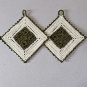 Op de afbeelding: Twee vierkante gehaakte pannenlappen. Elke pannenlap heeft een centraal olijfgroen ruitpatroon, een crèmekleurige rand en een bijpassende olijfgroene geschulpte rand. Er is een lus bevestigd om op te hangen. Handgemaakt.