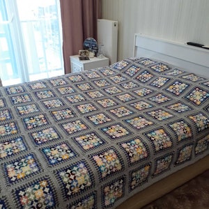 May include: A grey crocheted blanket with a colourful floral pattern. The blanket is on a bed with a white headboard.