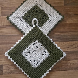 Può includere: Due presine all'uncinetto fatte a mano. Una è verde oliva con un motivo floreale bianco al centro, e l'altra è bianca con un motivo floreale verde oliva. Entrambe hanno una forma quadrata con un bordo smerlato e un anello per appenderle.