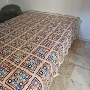 Può includere: Una coperta all'uncinetto fatta a mano, multicolore. La coperta presenta un motivo ripetuto di motivi quadrati in vari colori, tra cui rosso, crema, blu e viola. La coperta è drappeggiata su un letto, mostrando le sue dimensioni e il suo design intricato.