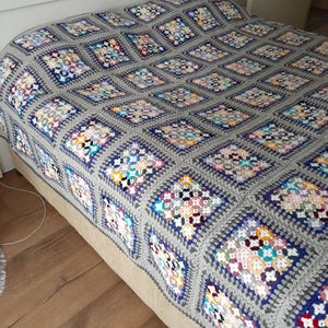 May include: A gray crocheted throw blanket with colorful squares featuring a floral pattern. The blanket is on a bed with a white headboard.