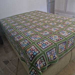 Puede incluir: Una manta de ganchillo hecha a mano y colorida con un borde verde. La manta presenta un diseño de patchwork de motivos cuadrados en varios colores, incluyendo azul, amarillo y morado. La manta está colocada sobre una cama.