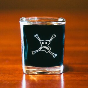 May include: A clear shot glass filled with a black liquid. The glass features a white skull and crossbones design with a frowning face.
