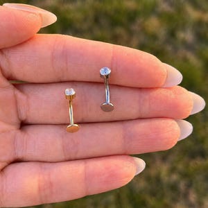 Op de afbeelding: Twee gebogen barbell-lichaamssieraden, één in goudkleur en één in zilverkleur, elk voorzien van een enkele heldere ronde edelsteen aan de bovenkant en een platte ronde schijfbasis aan de onderkant, tegen een wazige achtergrond.