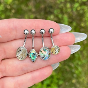 Puede incluir: Cuatro anillos de ombligo plateados con diferentes diseños de concha de abulón iridiscente. Los anillos están en una mano.