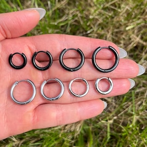 May include: A collection of eight hoop earrings arranged in two rows. The top row features four black hoop earrings of varying sizes, while the bottom row displays four silver-toned hoop earrings of varying sizes, all with a simple circular design.