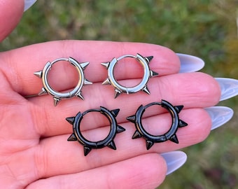 May include: Two pairs of hoop earrings, one pair in a polished silver tone and one pair in a matte black finish. Each circular hoop features a series of evenly spaced, pointed spikes protruding outward along the outer edge of the metal band.