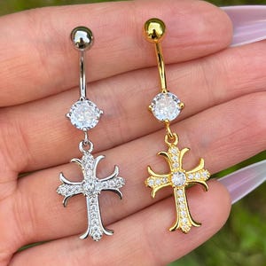 Puede incluir: Dos anillos de ombligo con un dije de cruz adornado con piedras. Un anillo es plateado con piedras transparentes y el otro es dorado con piedras transparentes.