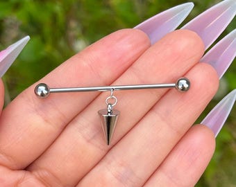 May include: A silver industrial barbell piercing with a dangling, cone-shaped charm. The barbell has a ball on each end. The charm is a polished, metallic cone. The jewelry is held against a blurred green background.