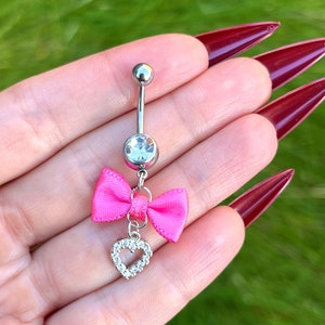 May include: A silver belly button ring with a clear crystal, a pink bow, and a dangling heart-shaped charm with clear crystals.