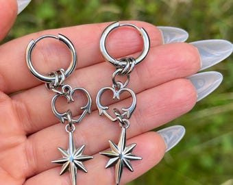 May include: A pair of silver hoop earrings. Each earring features a heart-shaped charm and a dangling starburst pendant. The earrings are displayed against a blurred green background.