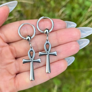 May include: A pair of silver-colored hoop earrings. Each earring features a small ball and a dangling ankh symbol. The ankh is a cross with a loop at the top, a symbol of life. The earrings are displayed against a blurred green background.