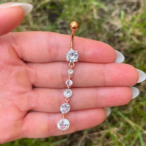 Op de afbeelding: Een roségoudkleurige navelpiercing met een verticaal hangend ontwerp. Het sieraad bestaat uit een bovenste balletje, een grotere ronde heldere steen en een bungelende ketting van vier steeds kleiner wordende ronde heldere stenen.