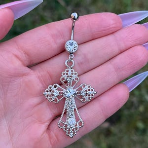 May include: A silver belly button ring with a dangling cross charm. The cross is adorned with clear rhinestones.
